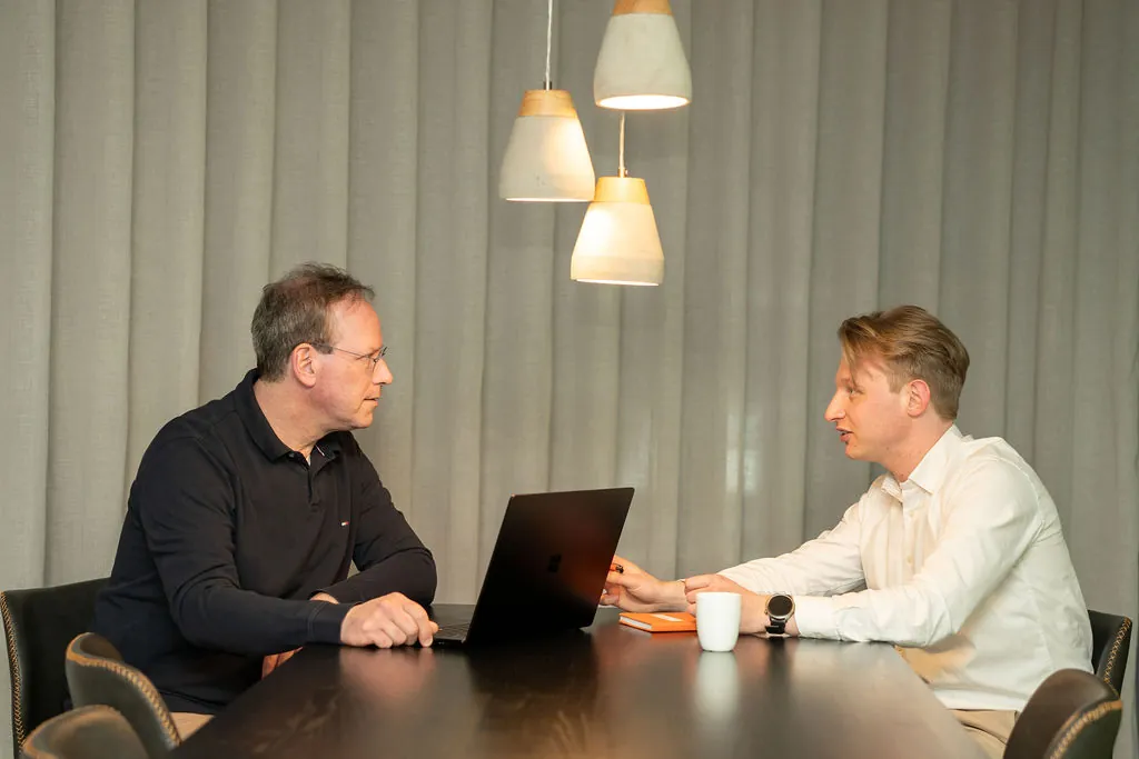 Een-op-een overleg aan tafel onder witte hanglampen, met een oranje notitieboek